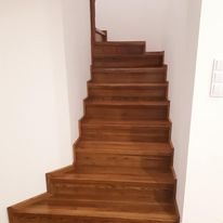 Brązowe, drewniane schody w jasnym wnętrzu, widok z dołu. Stopnie o wyraźnym usłojeniu, kontrastują z białą ścianą. Minimalistyczny design, brak poręczy.