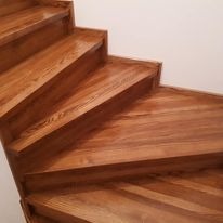 Polerowane, drewniane schody zabiegowe z widocznym usłojeniem, oświetlone z góry, z białym tłem ściany. Ujęcie z dołu, podkreślające detale wykończenia.