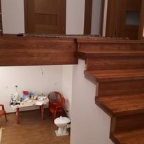 Drewniane schody z ciemnego drewna w domu w Sanoku. Widoczne stopnie i fragment konstrukcji. W tle pomieszczenie z meblami i akcesoriami. Ujęcie z dołu, perspektywa.