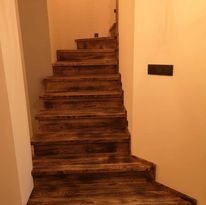 Rustykalne schody z ciemnego drewna z wyraźnym usłojeniem, oświetlone ciepłym światłem, prowadzące w górę. Widoczne detale wykończenia i kontrast z jasną ścianą.