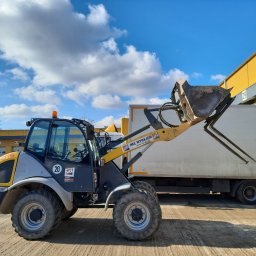 P&P - Żółto-szara ładowarka teleskopowa HKL na placu budowy, z podniesioną łyżką i widłami, na tle magazynu z rampami i błękitnego nieba z chmurami.