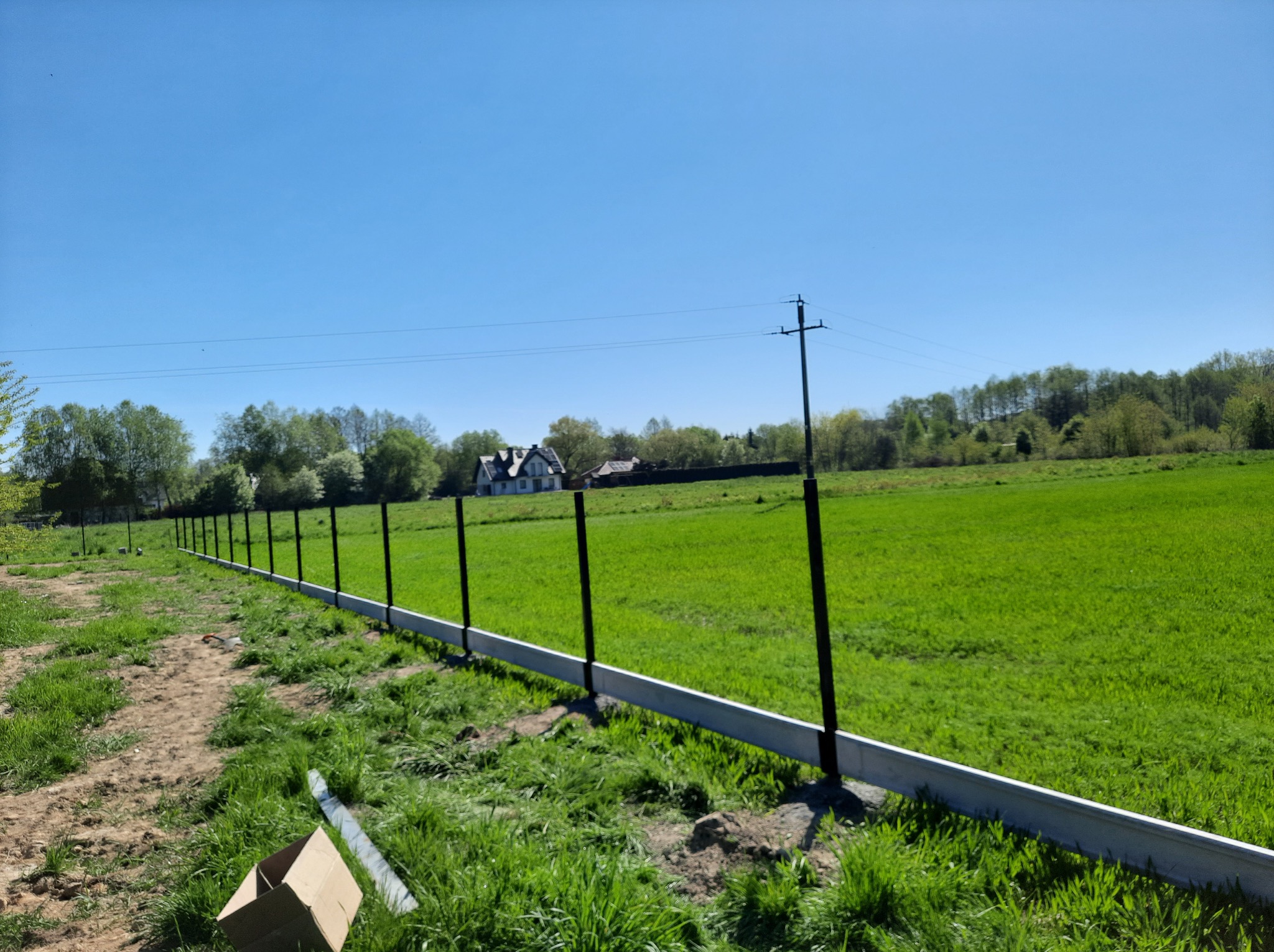 Montaż ogrodzenia panelowego na zielonej działce. Widoczne słupki i podmurówka, w tle dom i drzewa. Słoneczny dzień, błękitne niebo. Prace w toku, karton i element ogrodzenia na ziemi.
