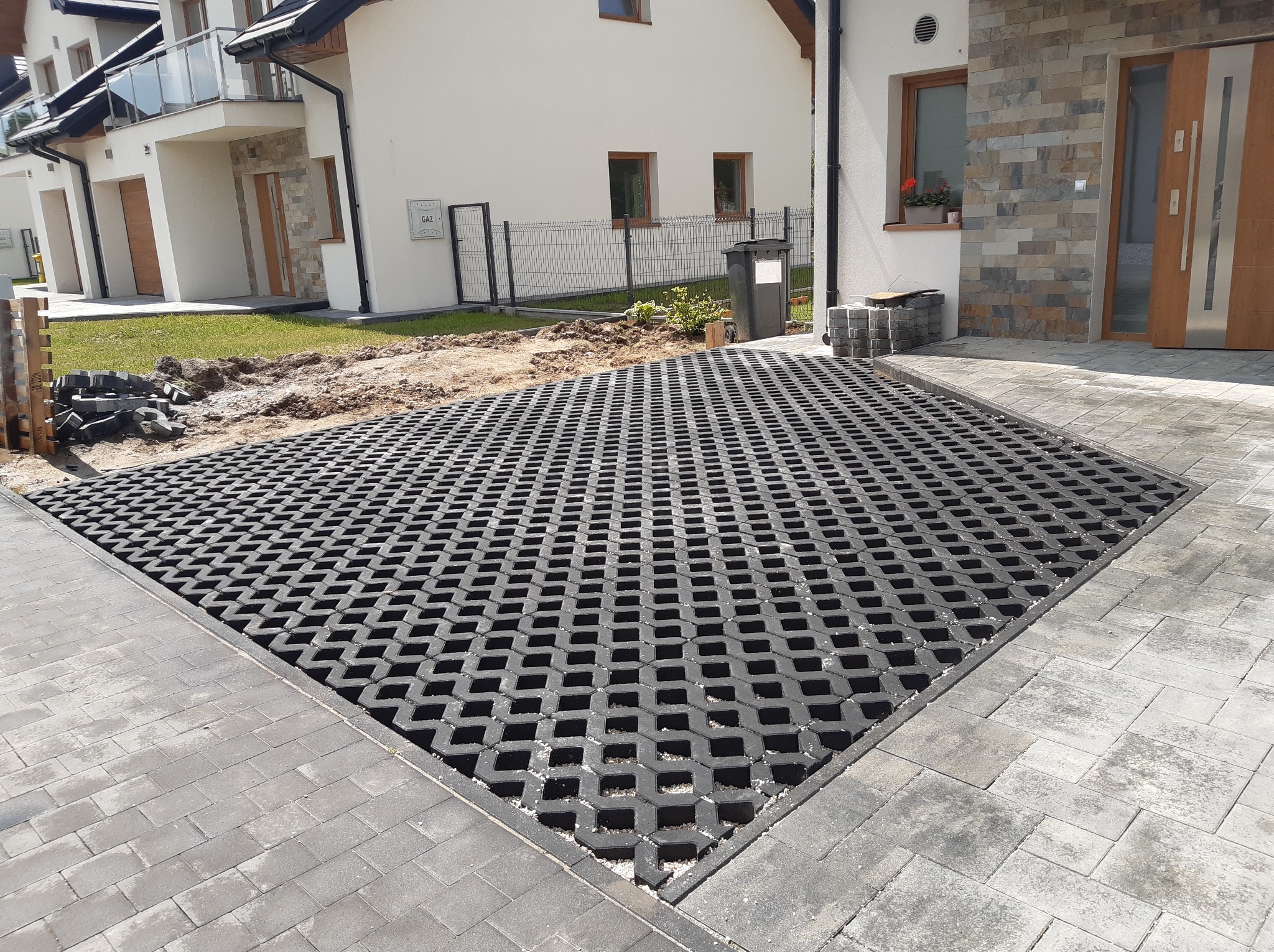 Ułożona, czarna ażurowa nawierzchnia na podjeździe do domu jednorodzinnego, obrzeża z szarej kostki brukowej. Częściowo widoczny trawnik i elewacja budynku.