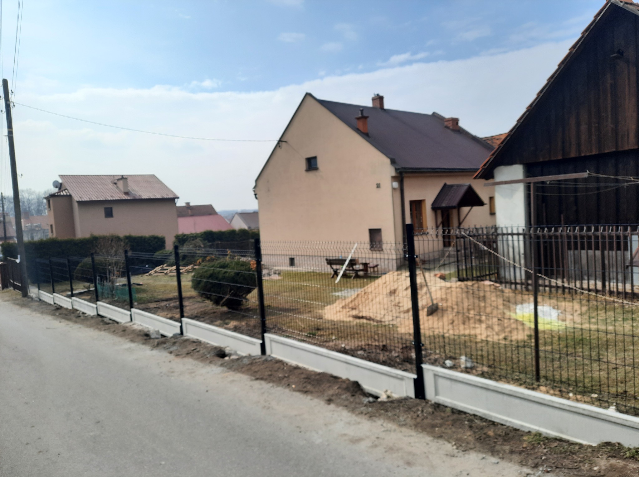 Nowoczesne ogrodzenie panelowe z podmurówką betonową, widok z ulicy. Na posesji widoczny piasek i narzędzia, sugerujące trwające prace wykończeniowe.
