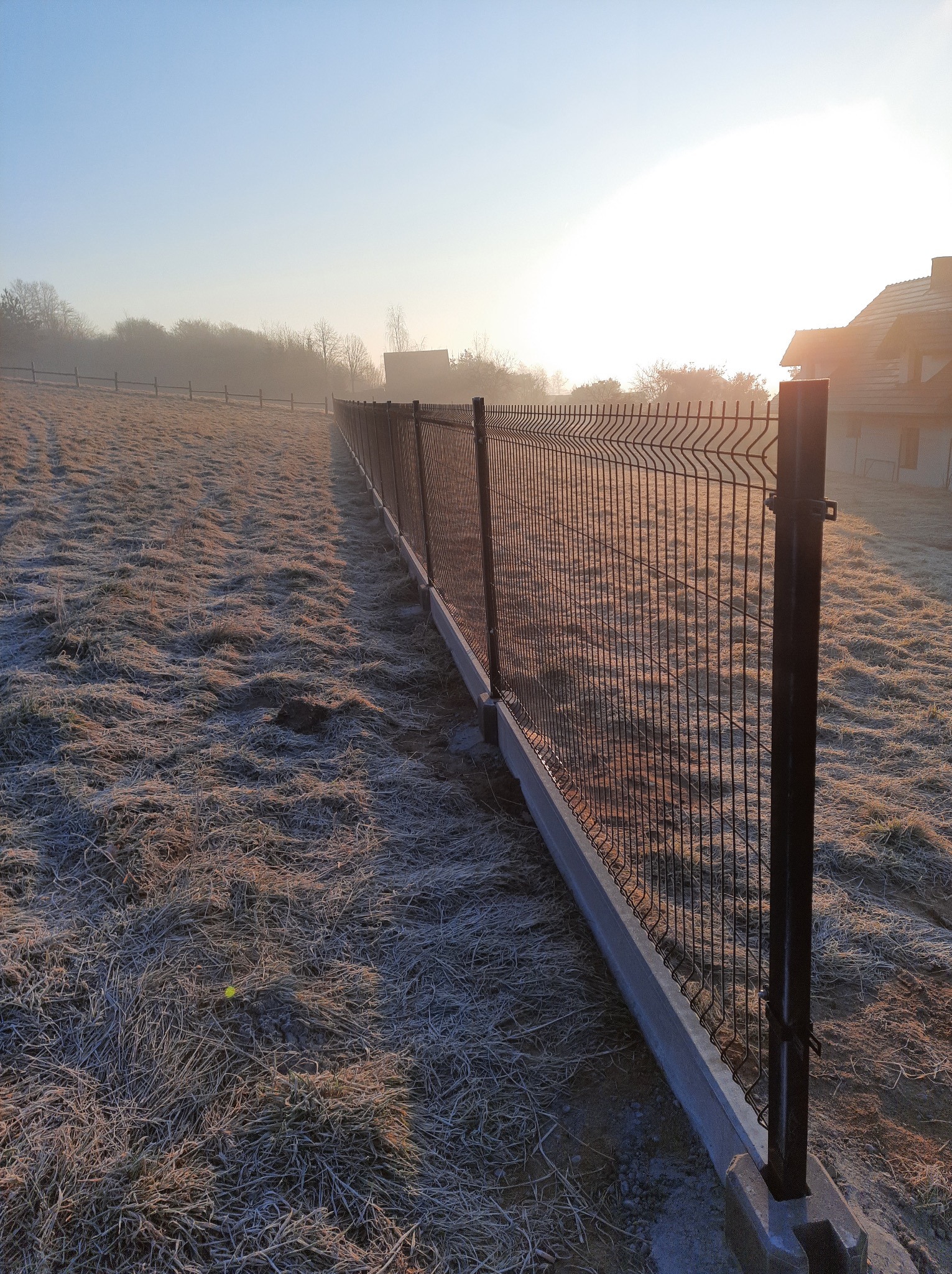 Nowoczesne ogrodzenie panelowe z podmurówką betonową, widok z perspektywy pola pokrytego szronem, w tle dom i drzewa w porannej mgle.