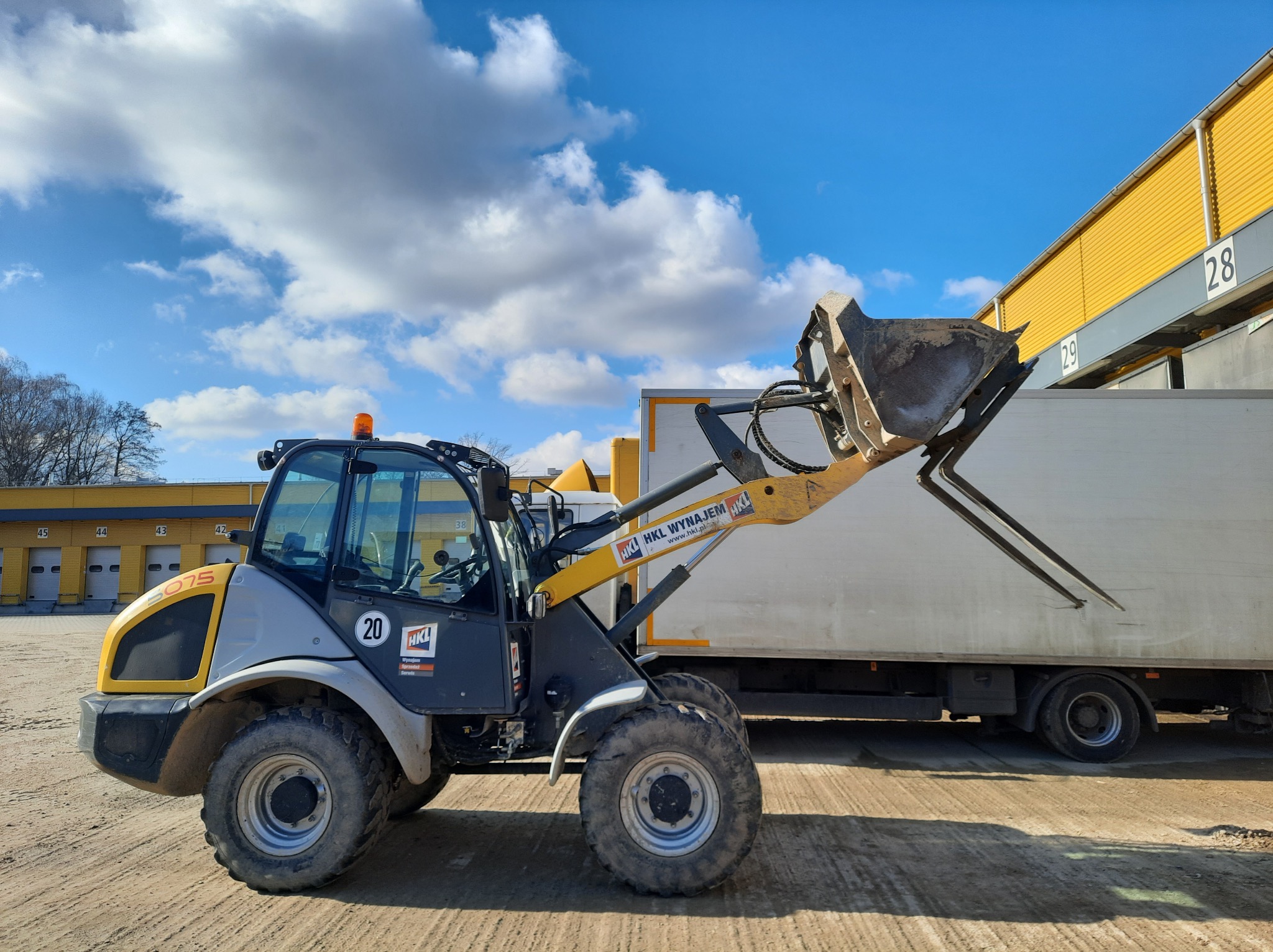Żółto-szara ładowarka teleskopowa HKL na placu budowy, z podniesioną łyżką i widłami, na tle magazynu z rampami i błękitnego nieba z chmurami.