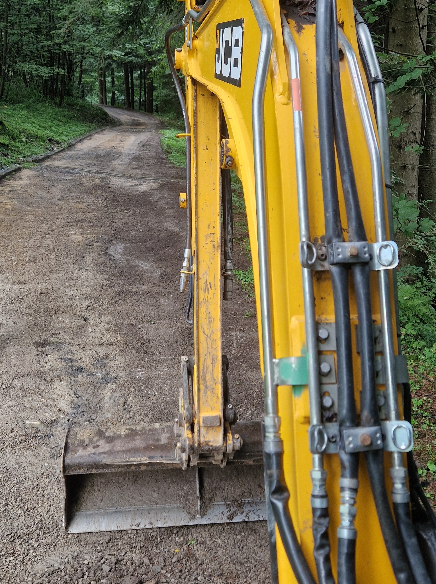 Żółta koparka JCB utwardza leśną drogę gruntową. Widoczne przewody hydrauliczne i lemiesz. W tle drzewa i zakręt drogi.