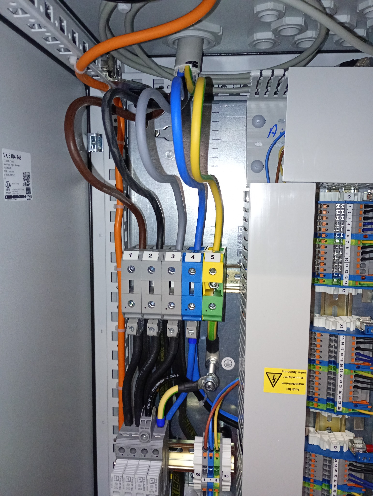 Rozdzielnica elektryczna z kolorowymi przewodami podłączonymi do listew zaciskowych. Widoczne oznaczenia numerów na złączach i ostrzeżenie o napięciu.