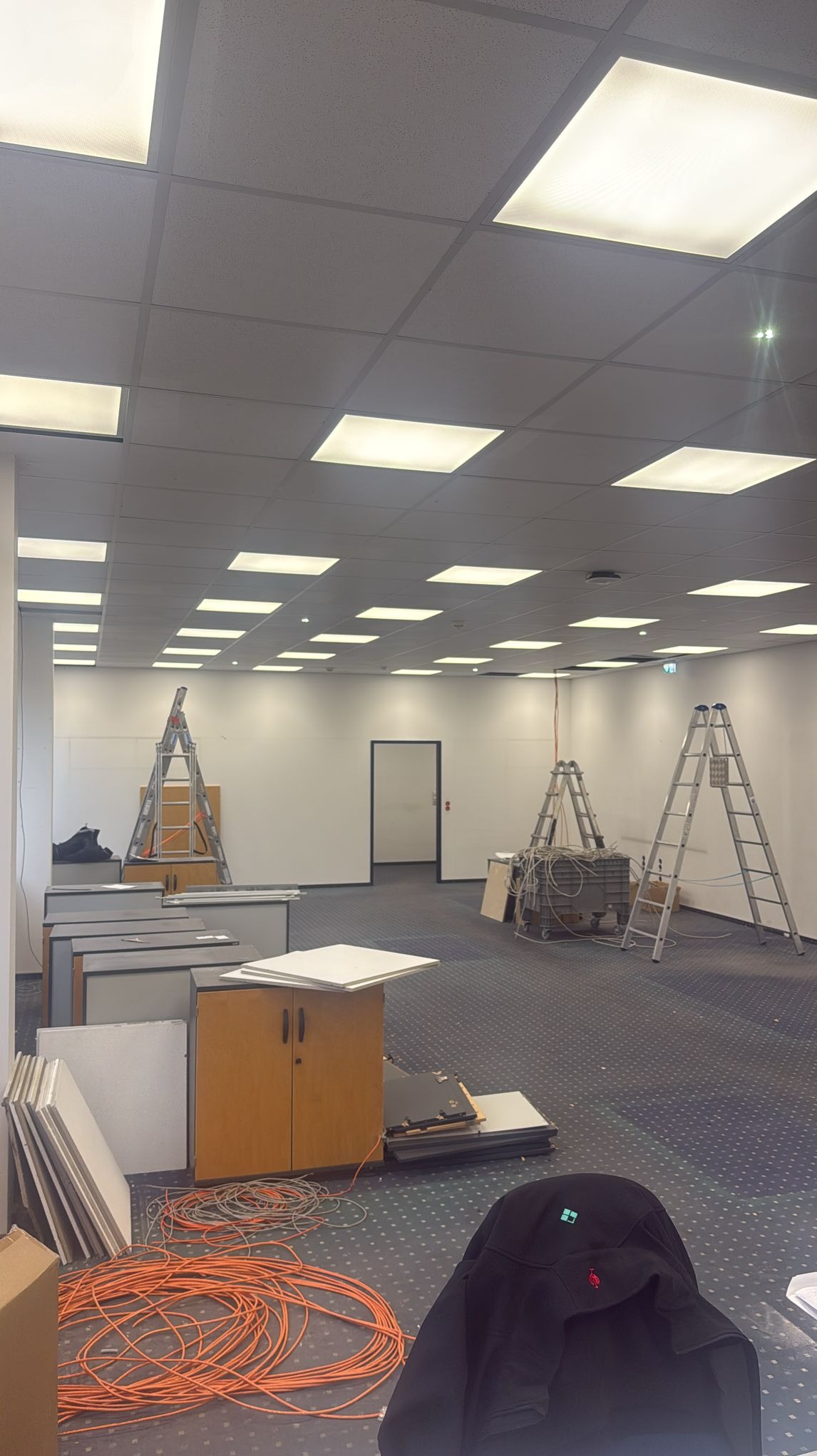 Montaż sufitu podwieszanego z oświetleniem rastrowym w biurze. Widoczne kable, drabiny i elementy konstrukcyjne. Prace w toku, przygotowanie do instalacji.