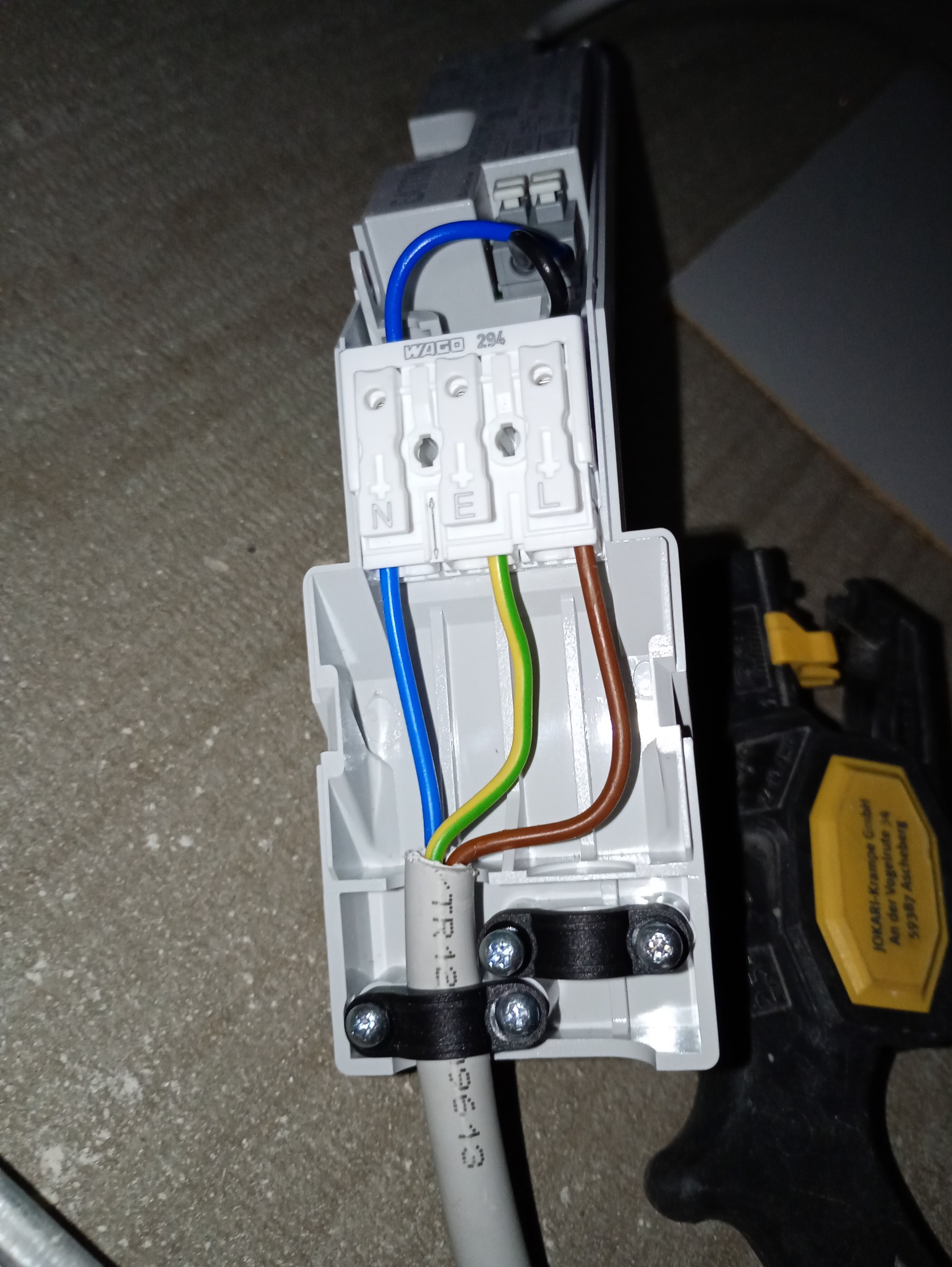 Podłączenie przewodów do listwy WAGO 294 z oznaczeniami N, E, L. Widoczne narzędzie do zdejmowania izolacji JOKARI. Kabel przymocowany uchwytami do podłoża.