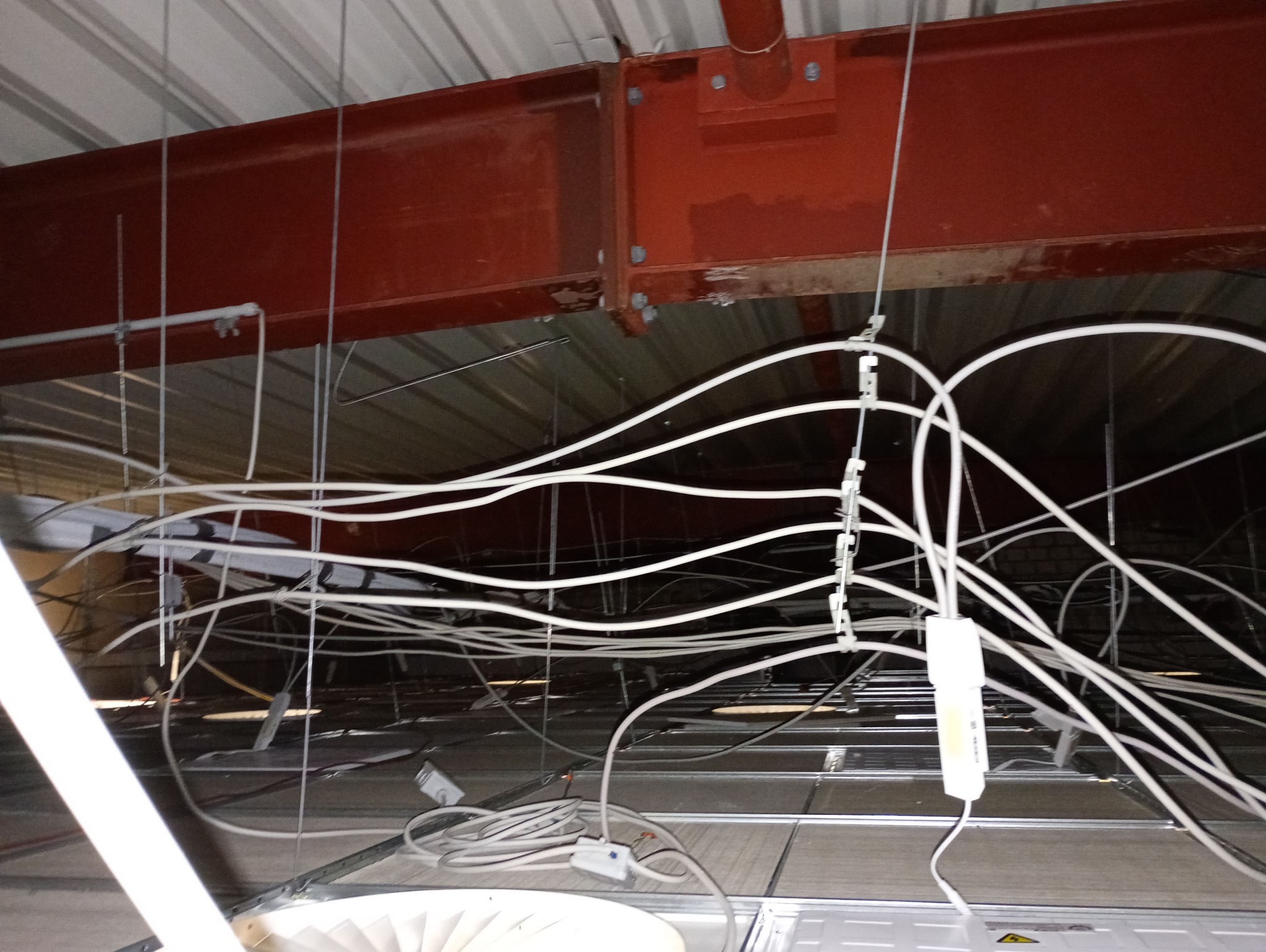 Skomplikowana sieć białych kabli pod sufitem podwieszanym, mocowanych do metalowej konstrukcji. Widoczne elementy oświetlenia i czerwona belka konstrukcyjna.