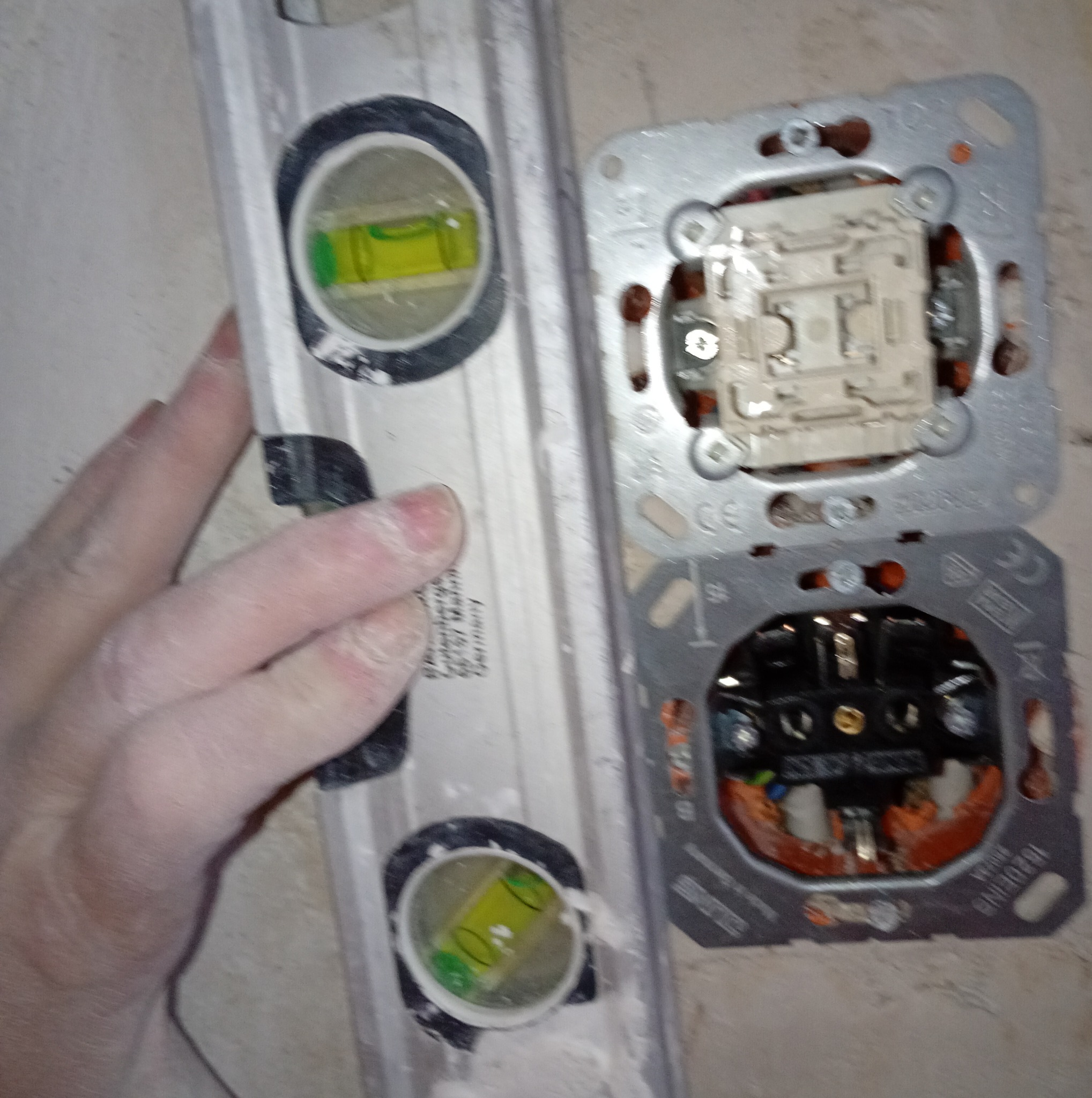 Poziomica przy gniazdku elektrycznym w trakcie montażu. Widoczne przewody i metalowa obudowa gniazdka. Ręka przytrzymuje poziomicę, sprawdzając poziom.