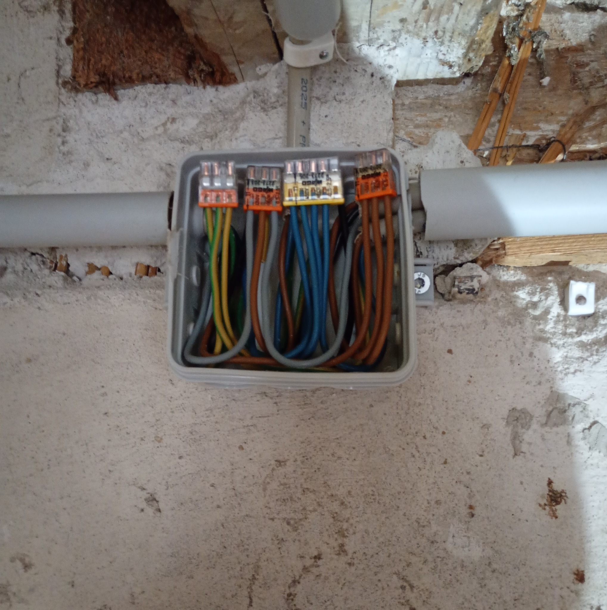 Szara puszka elektryczna z podłączonymi przewodami w kolorach: zielonym, żółtym, brązowym i niebieskim. Widoczne złącza WAGO. Tło stanowi surowa ściana i fragment instalacji.