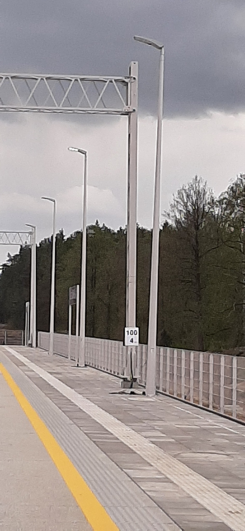 Nowoczesne oświetlenie LED na peronie kolejowym z szarymi słupami i metalową konstrukcją nad torami, w tle zieleń drzew i pochmurne niebo.