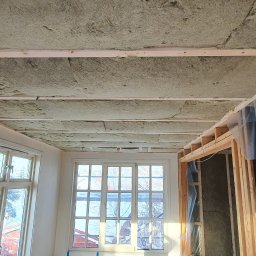 Wiercimy Temat - Poddasze z widoczną izolacją z wełny mineralnej między drewnianymi belkami stropowymi. W tle okna i fragment ściany z niedokończonym wykończeniem.