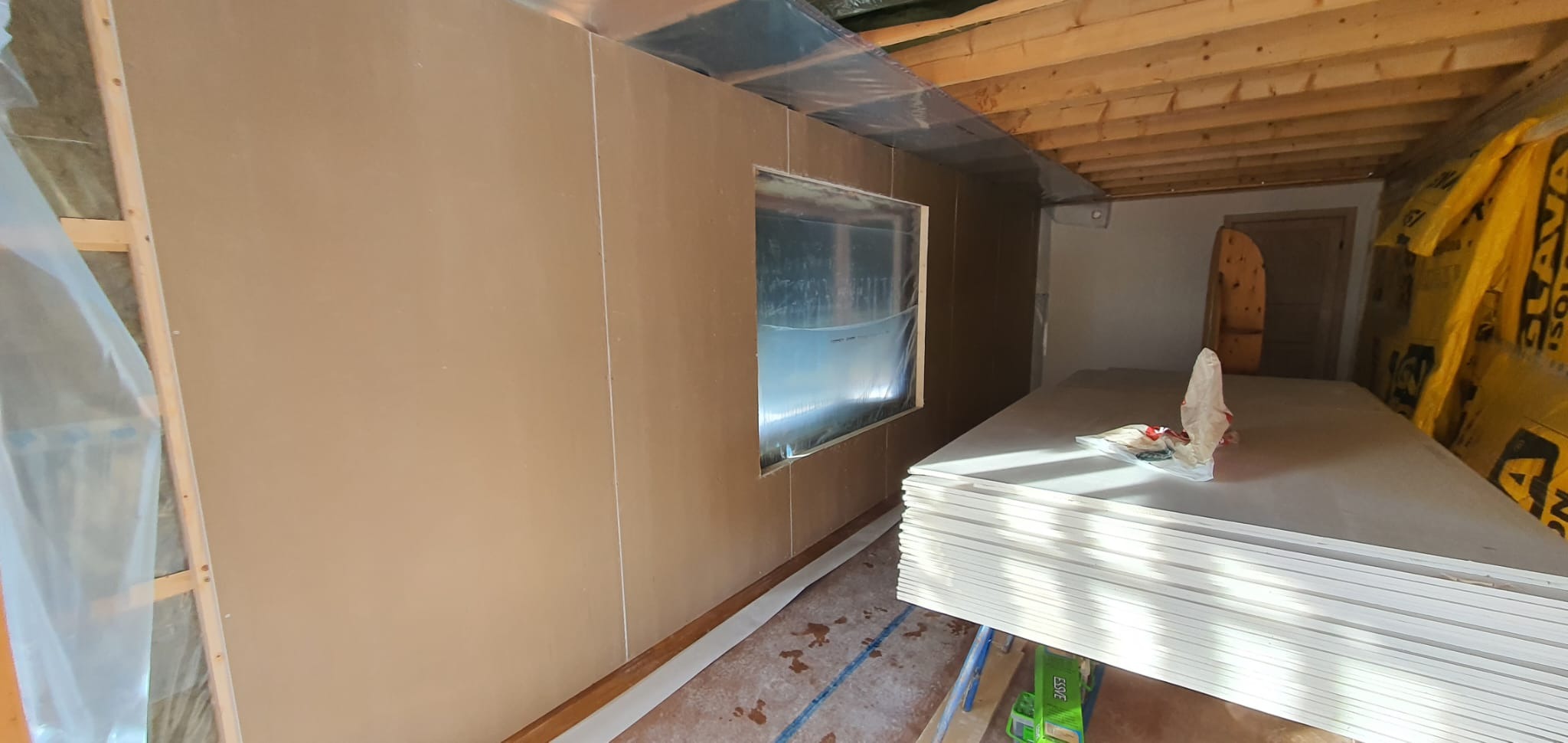 Wnętrze w trakcie prac wykończeniowych: ściany obłożone płytami, widoczne ocieplenie, stos płyt na podłodze. Surowy stan, widoczne elementy konstrukcyjne.