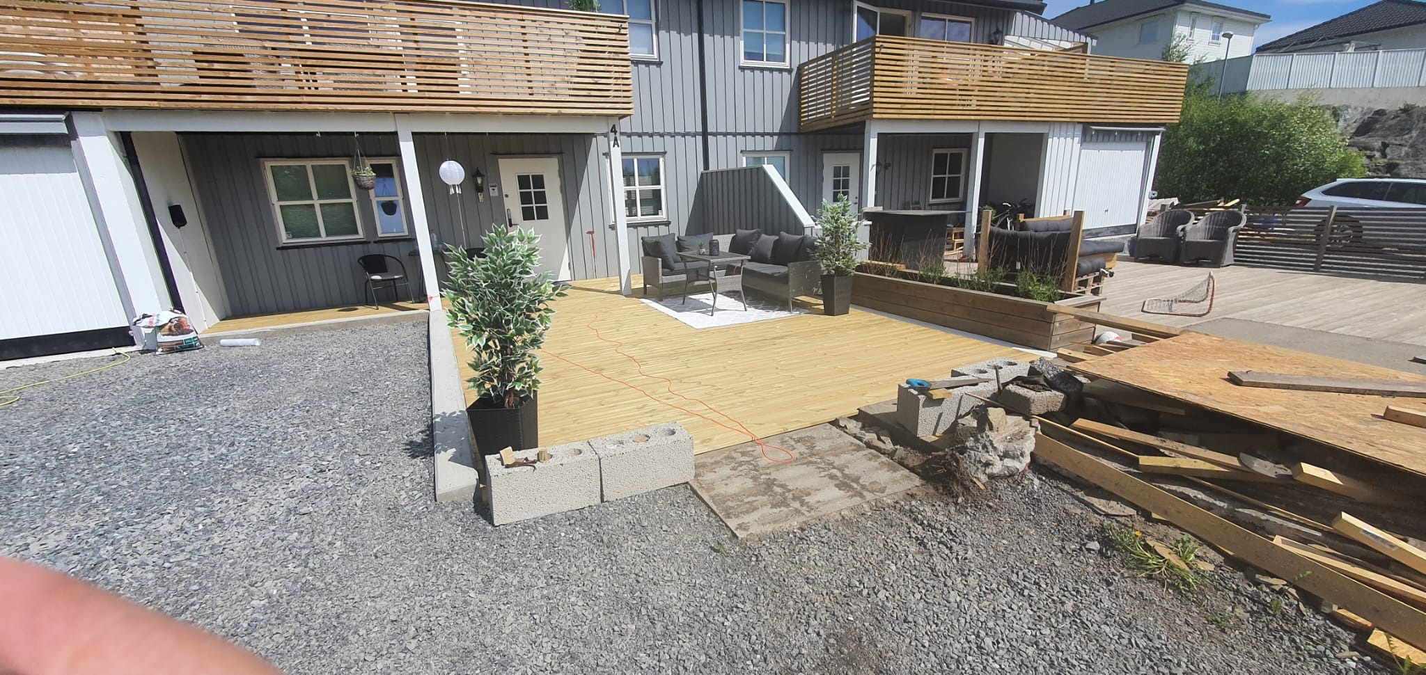 Drewniany taras przy szarym domu z balkonami, częściowo w trakcie budowy. Widoczne meble ogrodowe, rośliny w donicach i materiały budowlane.