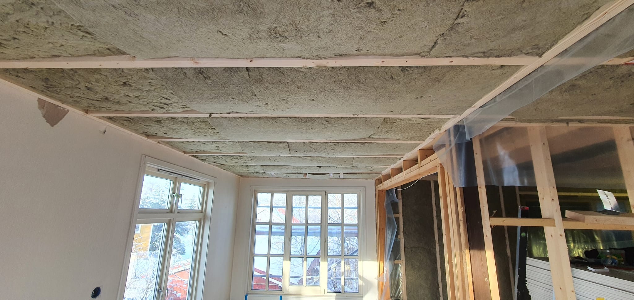 Poddasze z widoczną izolacją z wełny mineralnej między drewnianymi belkami stropowymi. W tle okna i fragment ściany z niedokończonym wykończeniem.