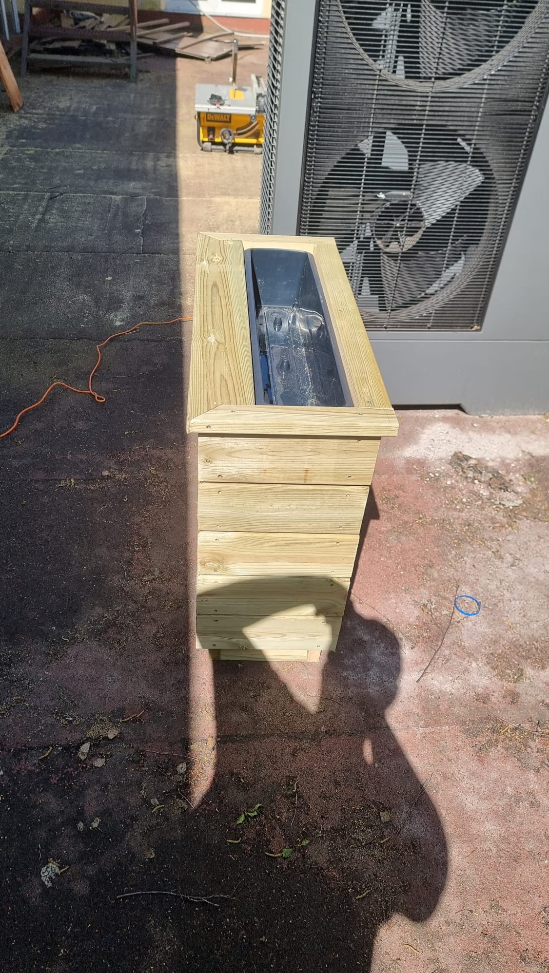 Drewniana donica na zewnątrz, wykonana na zamówienie. Widoczna faktura drewna i metalowy wkład. W tle narzędzie do cięcia drewna i klimatyzator.