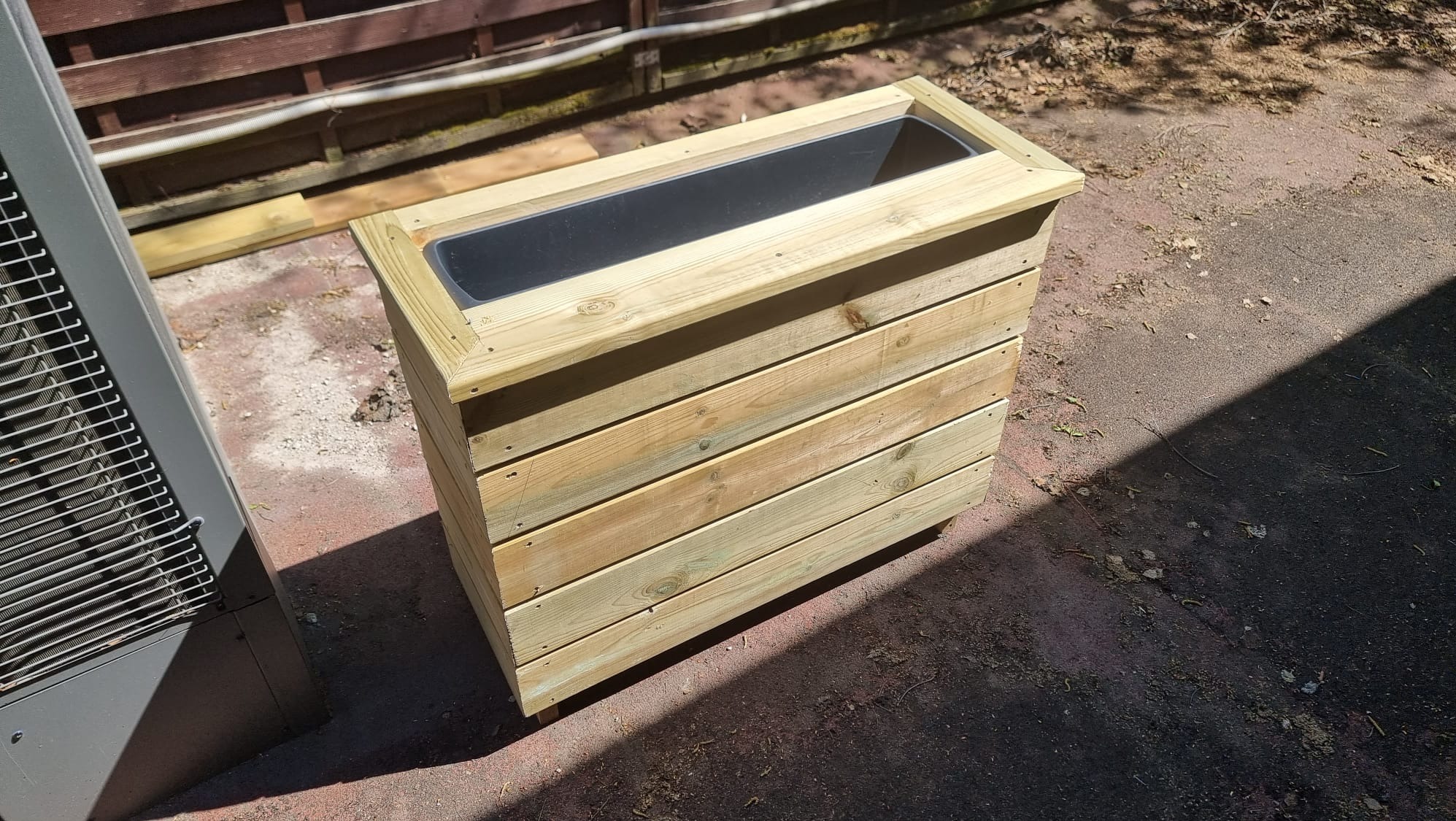 Drewniana donica na zamówienie, wykonana z jasnych desek, z czarnym wkładem. Widoczne łączenia śrubami. Obok stoi szara jednostka zewnętrzna klimatyzacji.