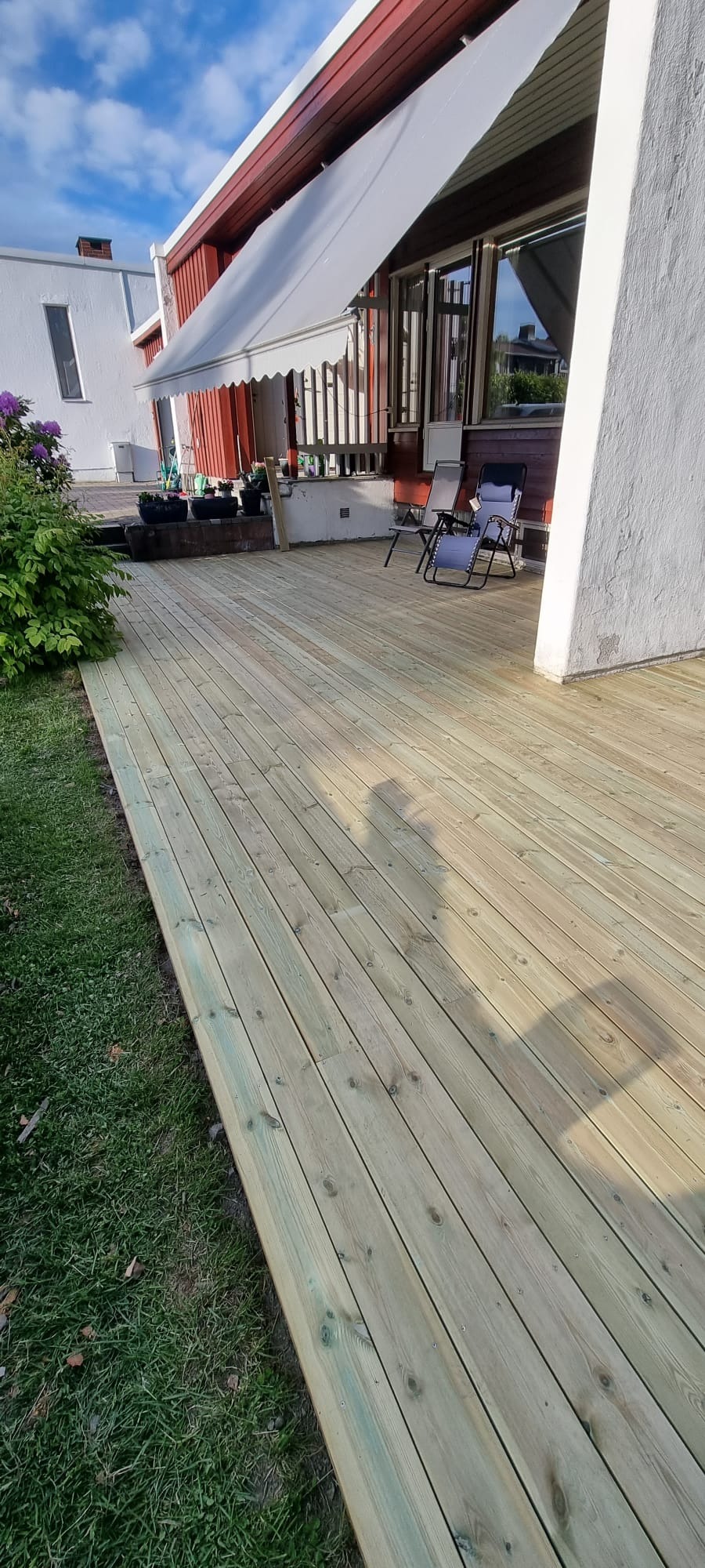 Nowa, jasna deska tarasowa z widocznymi śladami montażu, przylegająca do domu z markizą. Dwa krzesła ogrodowe w tle, cień osoby na deskach.