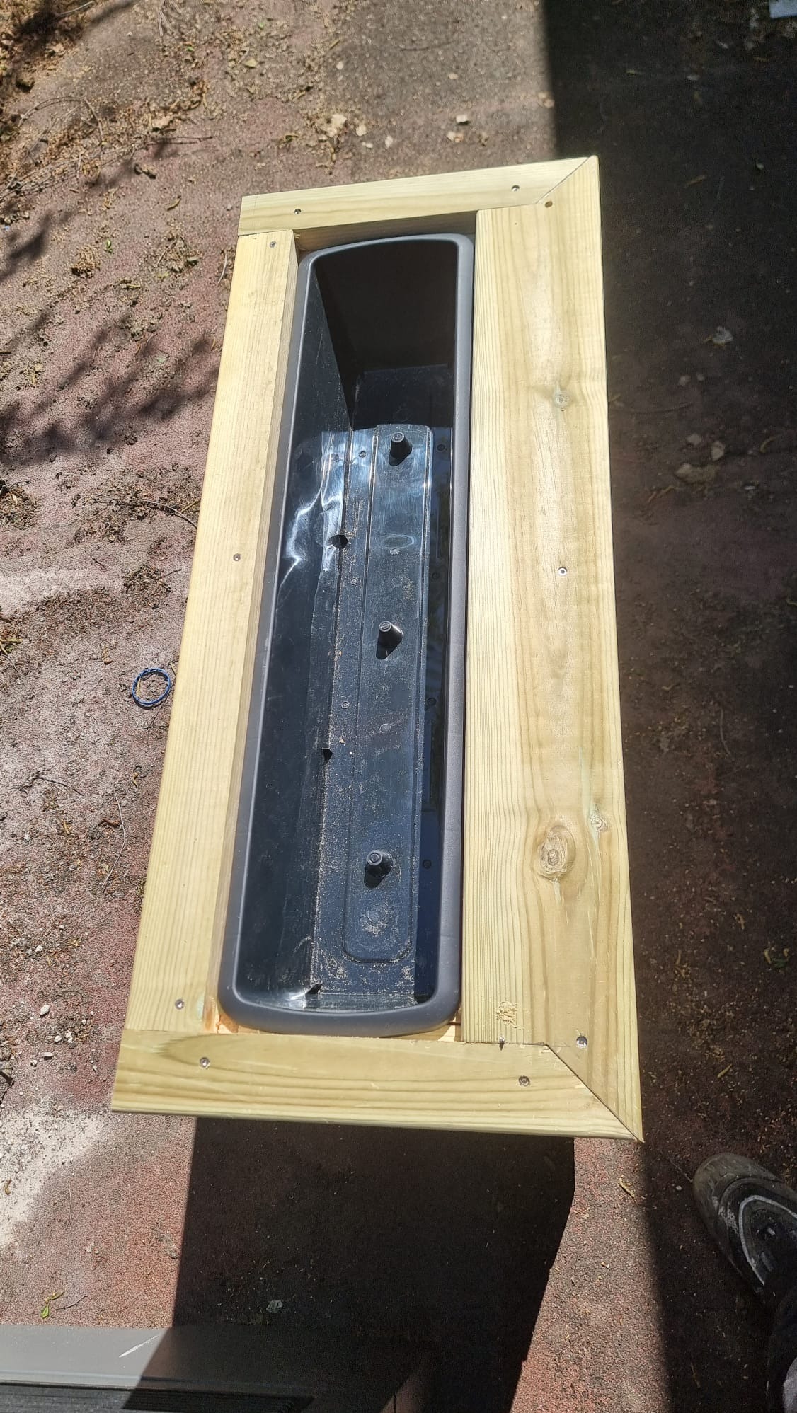 Drewniana donica na wymiar z czarnym wkładem, skręcona śrubami, widoczna z góry na zewnątrz. Jasne drewno, precyzyjne łączenia, cień na podłożu.