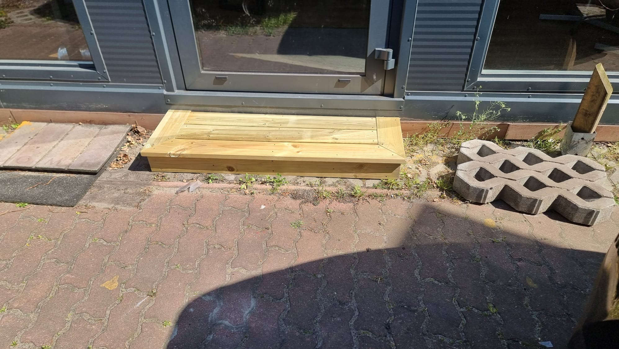 Drewniany podest wejściowy do pawilonu w Świnoujściu. Nowy, jasny, z desek, zamontowany przed szarymi drzwiami. Kostka brukowa i elementy betonowe obok.