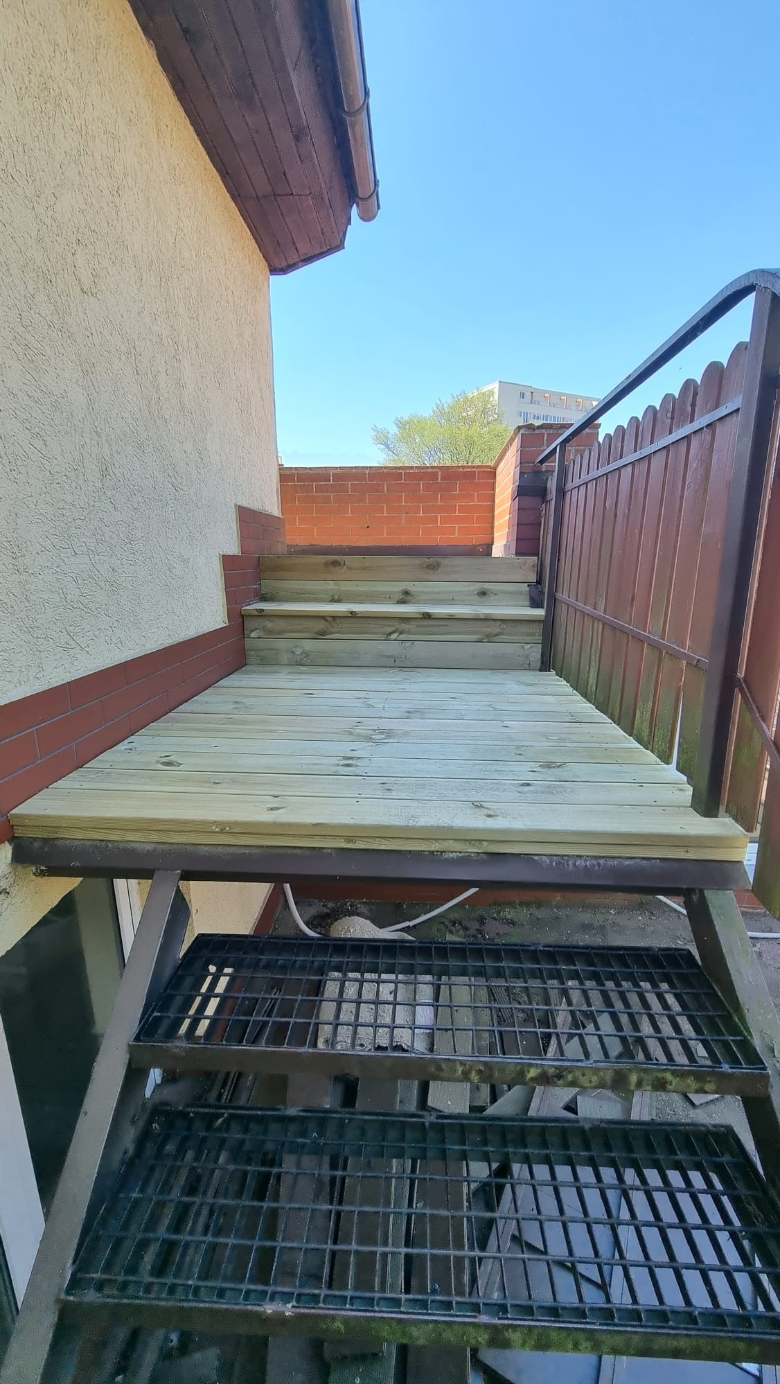 Drewniane schody wejściowe z metalową konstrukcją, prowadzące do budynku z ceglanym wykończeniem. Widok z dołu, błękitne niebo w tle.