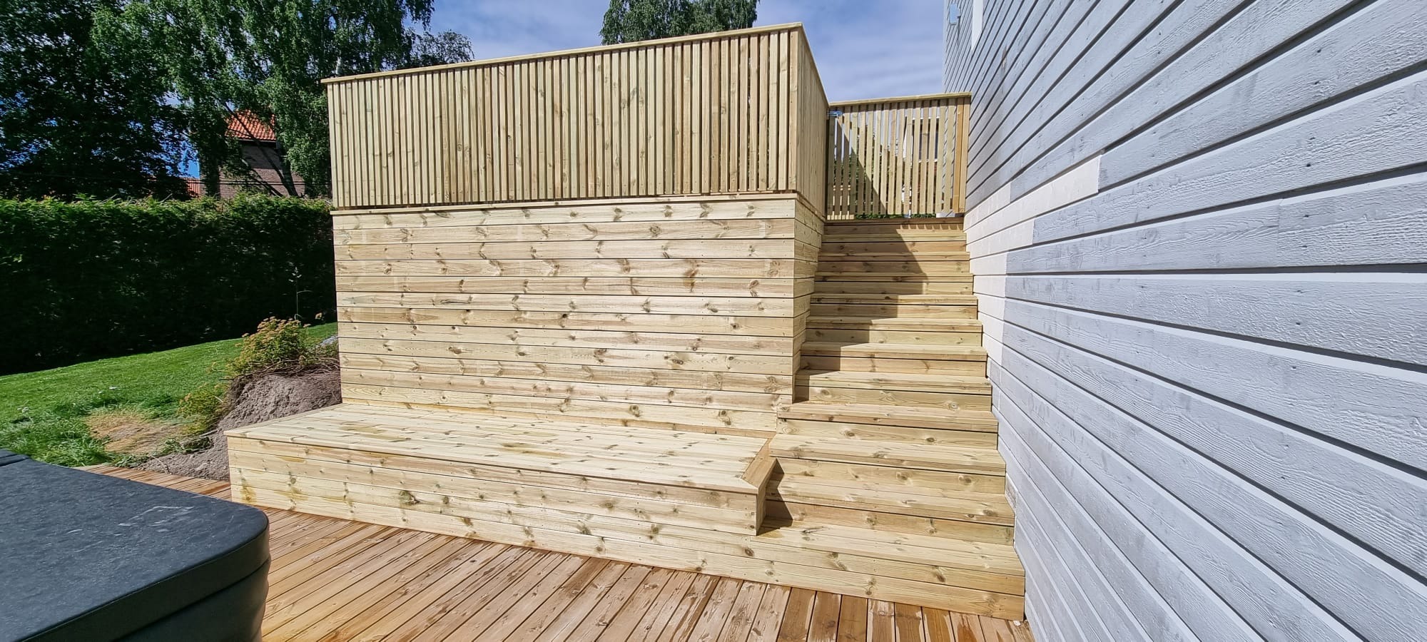Drewniany, dwupoziomowy taras z barierkami i schodami, przylegający do jasnego domu z drewnianą elewacją. Widoczne przygotowane miejsce pod basen.