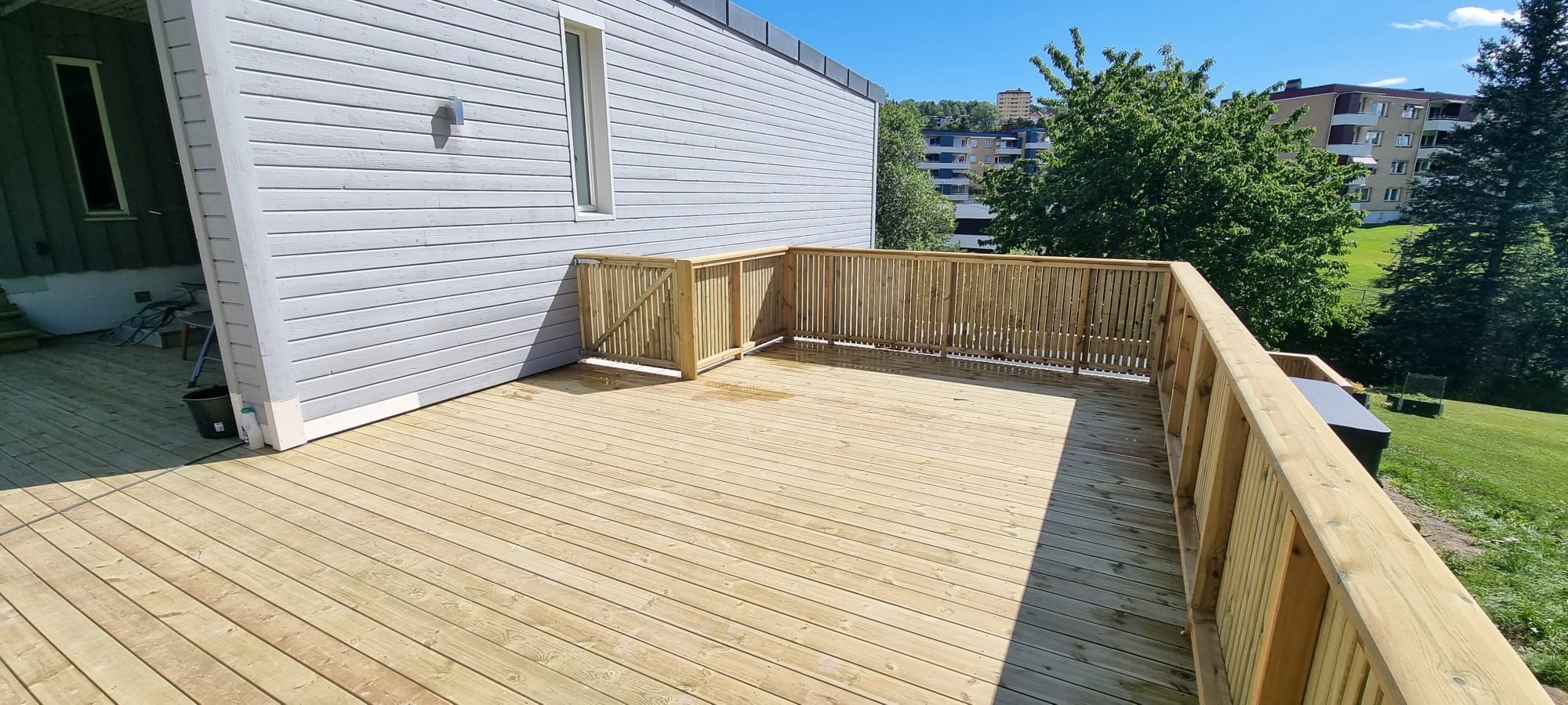 Jasny, drewniany taras z barierkami przy szarym domu z sidingu. W tle zieleń drzew i budynki mieszkalne. Słoneczny dzień.
