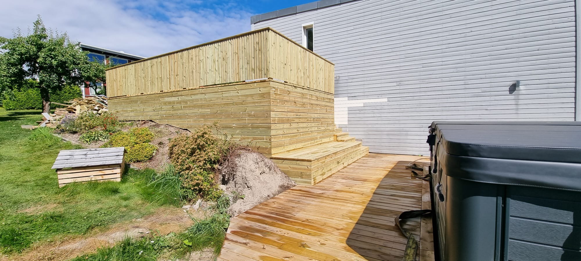 Dwupoziomowy taras z jasnego drewna z barierkami i schodami, przylegający do domu z szarą elewacją. Obok widoczna wanna z hydromasażem i fragment ogrodu.