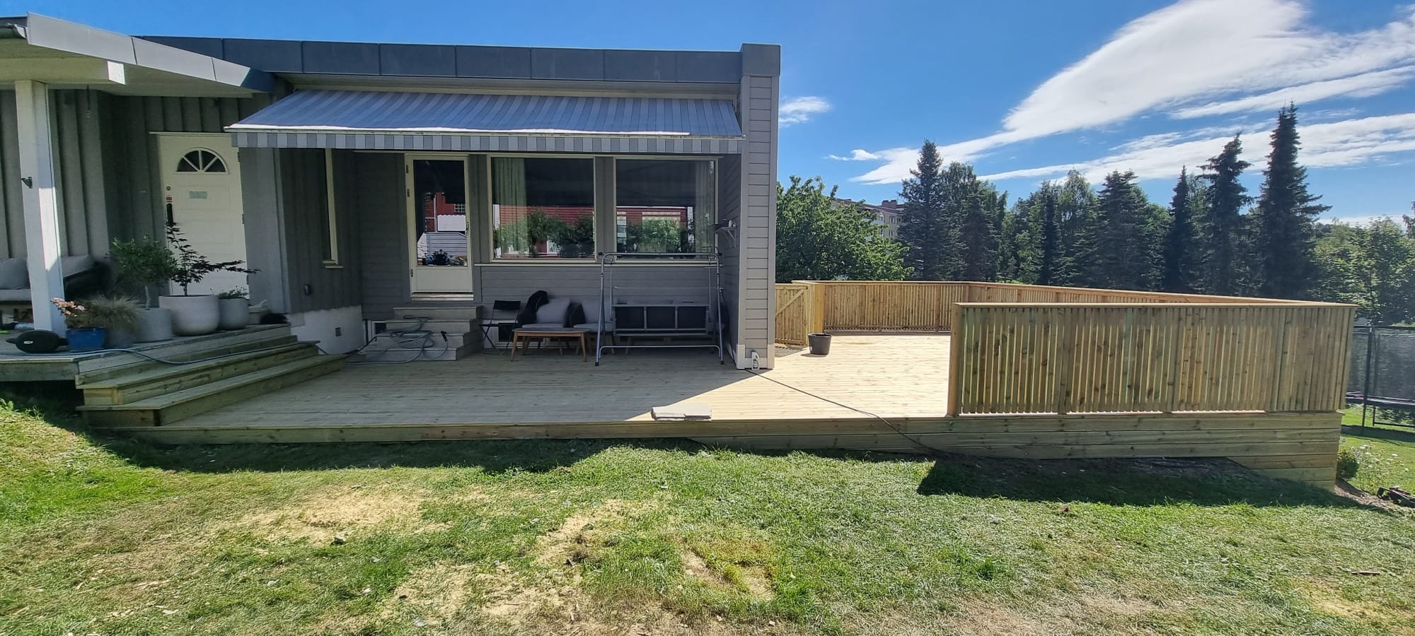 Dwupoziomowy taras z jasnego drewna z barierkami, przylegający do szarego domu z markizą. Widoczne schody, meble ogrodowe i przygotowane miejsce pod basen.