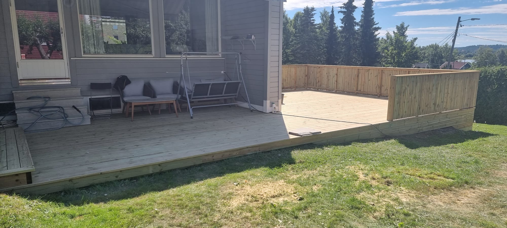 Dwupoziomowy taras z drewnianą balustradą, meblami ogrodowymi i przygotowanym miejscem pod basen, przylegający do domu z szarą elewacją.