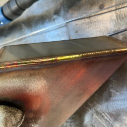 Metal Idustry Company Sp z O.O. - Metalowy element po spawaniu z widocznymi kolorami spoiny, leżący na metalowym stole warsztatowym. Widoczny fragment rękawicy ochronnej.