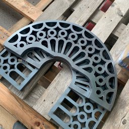 Metal Idustry Company Sp z O.O. - Wycięte laserowo, metalowe elementy dekoracyjne z powtarzalnym wzorem geometrycznym, ułożone na drewnianej palecie. Widoczny fragment buta dla skali.