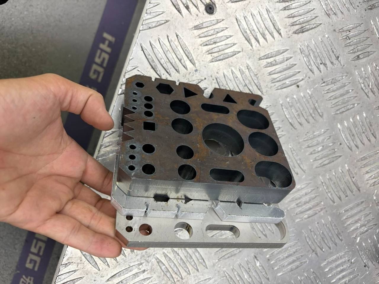 Metalowy blok z otworami o różnych kształtach, trzymany w dłoni na tle blachy ryflowanej. Precyzyjna obróbka metalu, widoczne detale i różnorodność kształtów.
