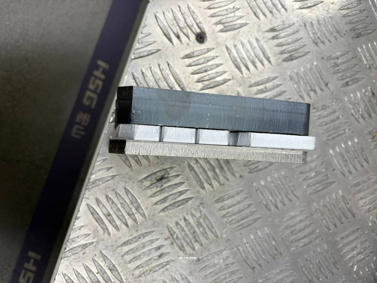 Precyzyjnie ułożone, różnorodne profile metalowe na tle blachy ryflowanej. Widoczne detale obróbki i materiałów, w tym element z napisem HSG.