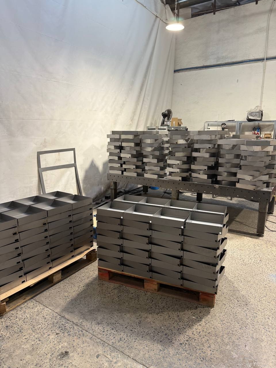 Stosy metalowych, szarych pojemników na paletach w hali produkcyjnej. Widoczne spawane konstrukcje i narzędzia w tle. Oświetlenie górne.