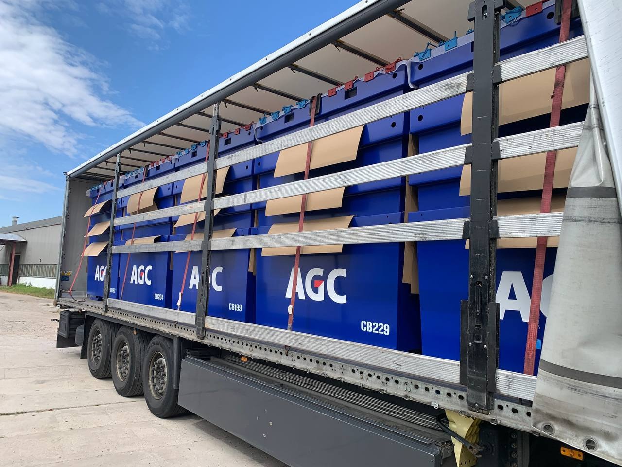 Ciężarówka z otwartą naczepą, załadowana niebieskimi, metalowymi kontenerami z logo AGC, zabezpieczonymi pasami i kartonem. Widok z boku, w tle budynek.