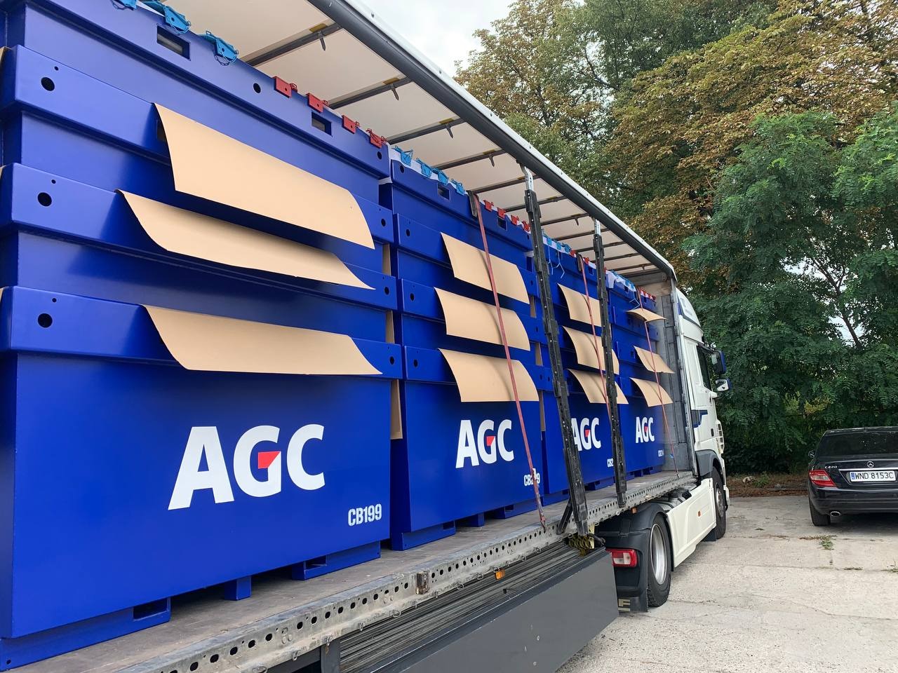 Transport niebieskich, metalowych konstrukcji z logo AGC na naczepie ciężarówki, zabezpieczonych kartonem. Widoczne drzewa w tle i zaparkowany samochód osobowy.