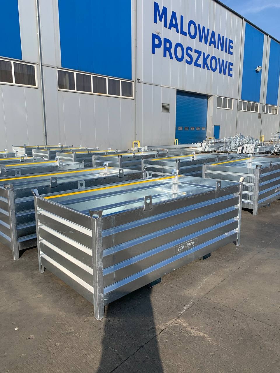 Stalowe, ocynkowane pojemniki z żółtymi akcentami, ustawione rzędami przed budynkiem z napisem 'Malowanie Proszkowe'.