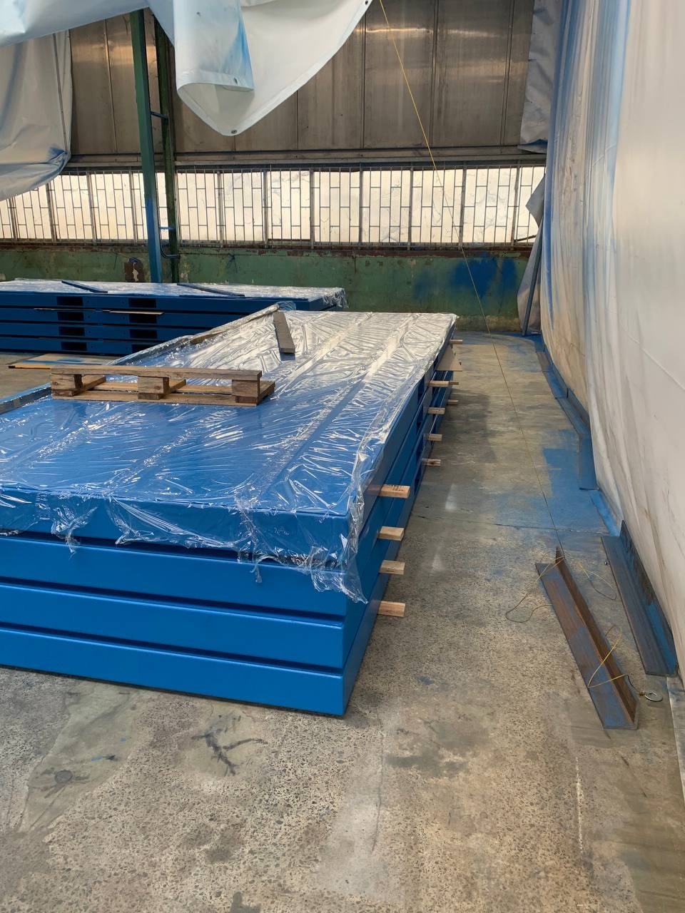 Stos niebieskich, metalowych elementów, zabezpieczonych folią, na drewnianej palecie w hali produkcyjnej. Widoczne okna i konstrukcja dachu.