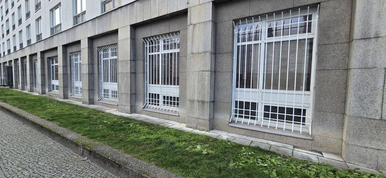 Białe, metalowe kraty okienne zabezpieczające okna w szarym, kamiennym budynku. Widok z zewnątrz, z perspektywy trawnika. Ochrona antywłamaniowa.