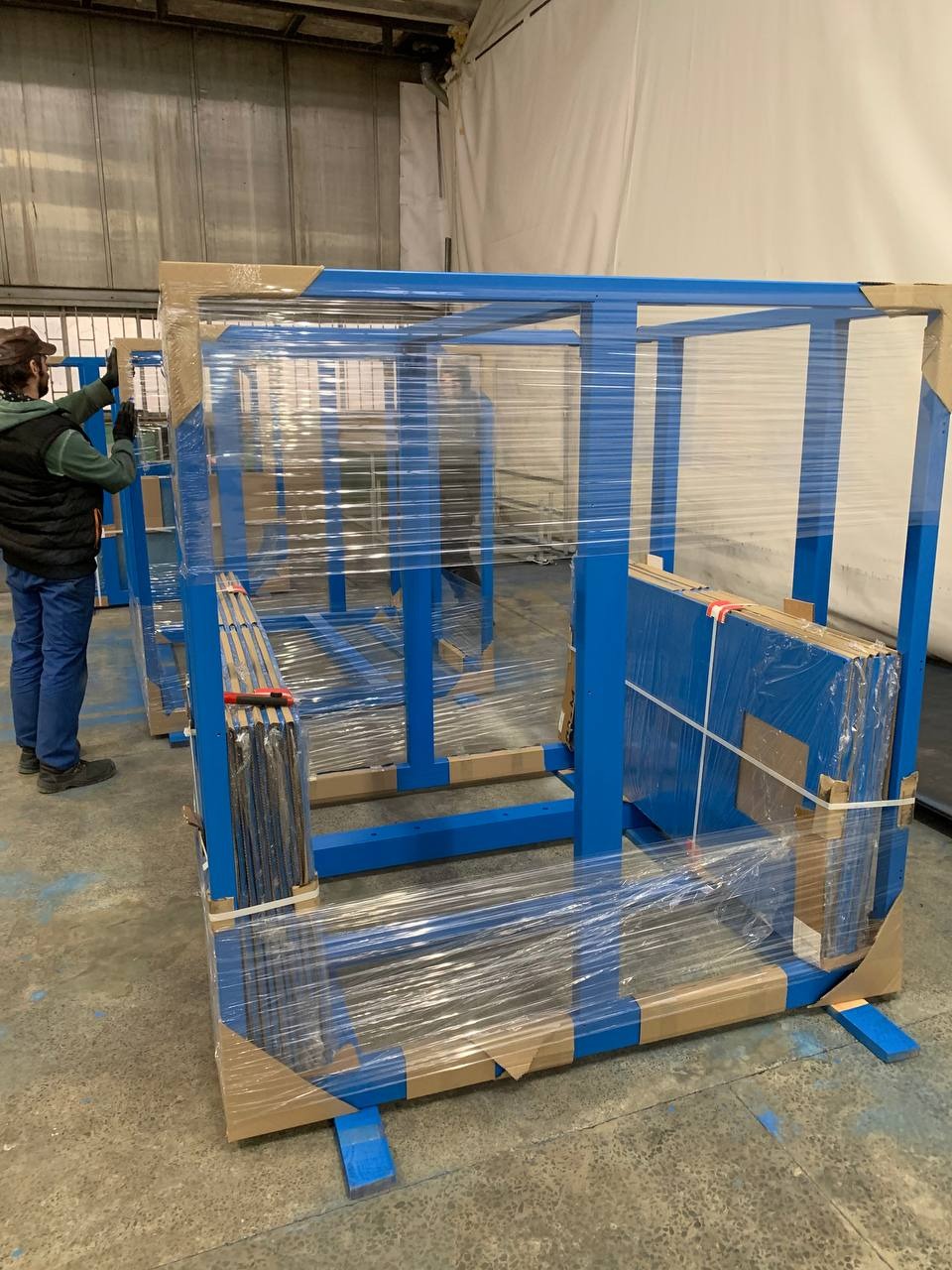 Niebieskie, metalowe konstrukcje w folii stretch, zabezpieczone kartonem, przygotowane do transportu. Widoczny pracownik w tle.