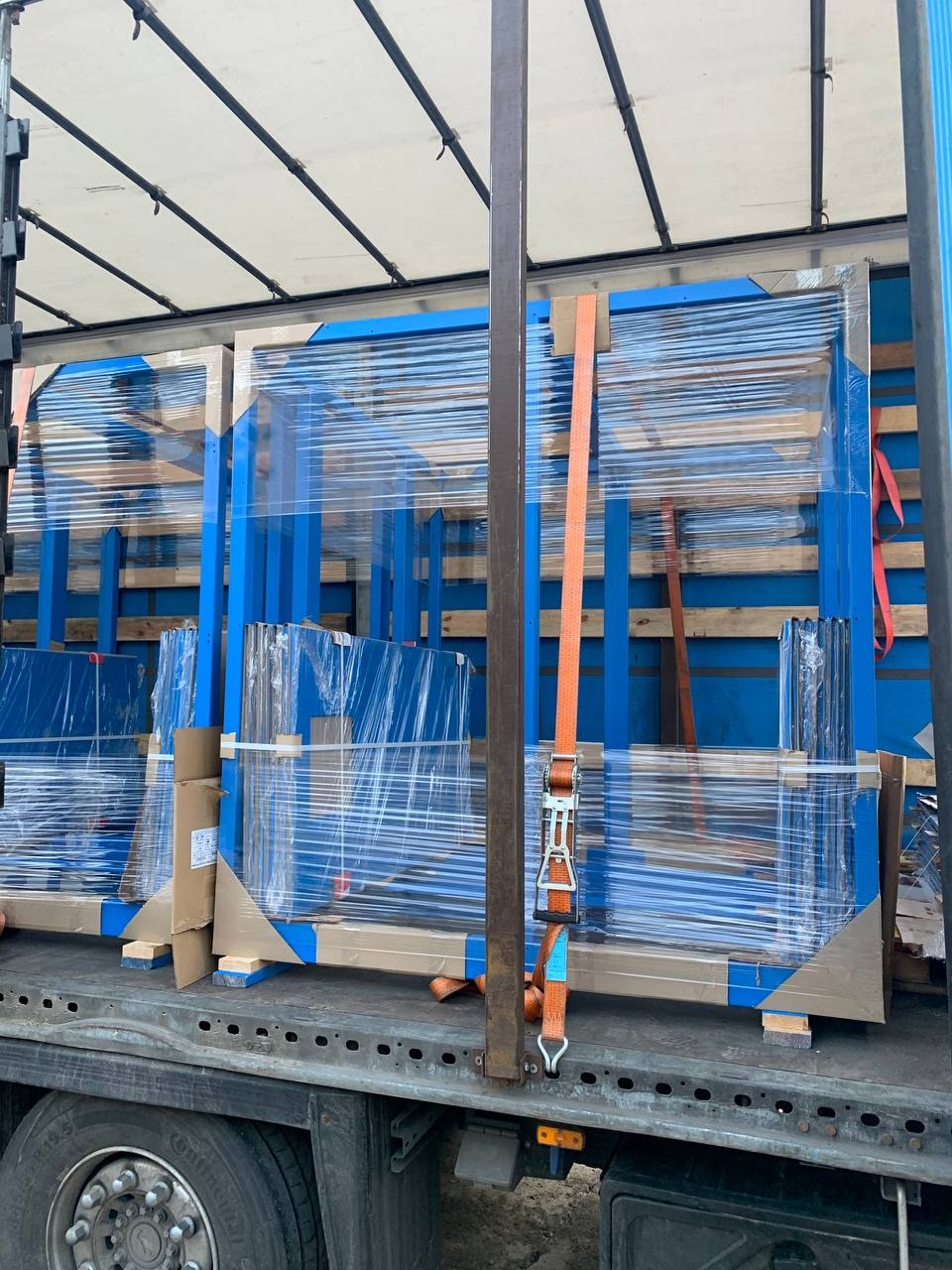 Transport niebieskich, metalowych konstrukcji w folii stretch, zabezpieczonych pasami transportowymi wewnątrz naczepy. Widoczne elementy drewniane i kartonowe.