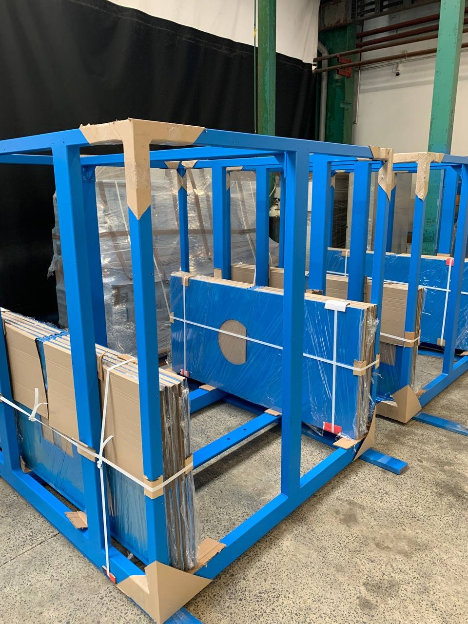 Niebieska, metalowa konstrukcja transportowa z zapakowanymi elementami, zabezpieczonymi kartonem i folią, w hali produkcyjnej.