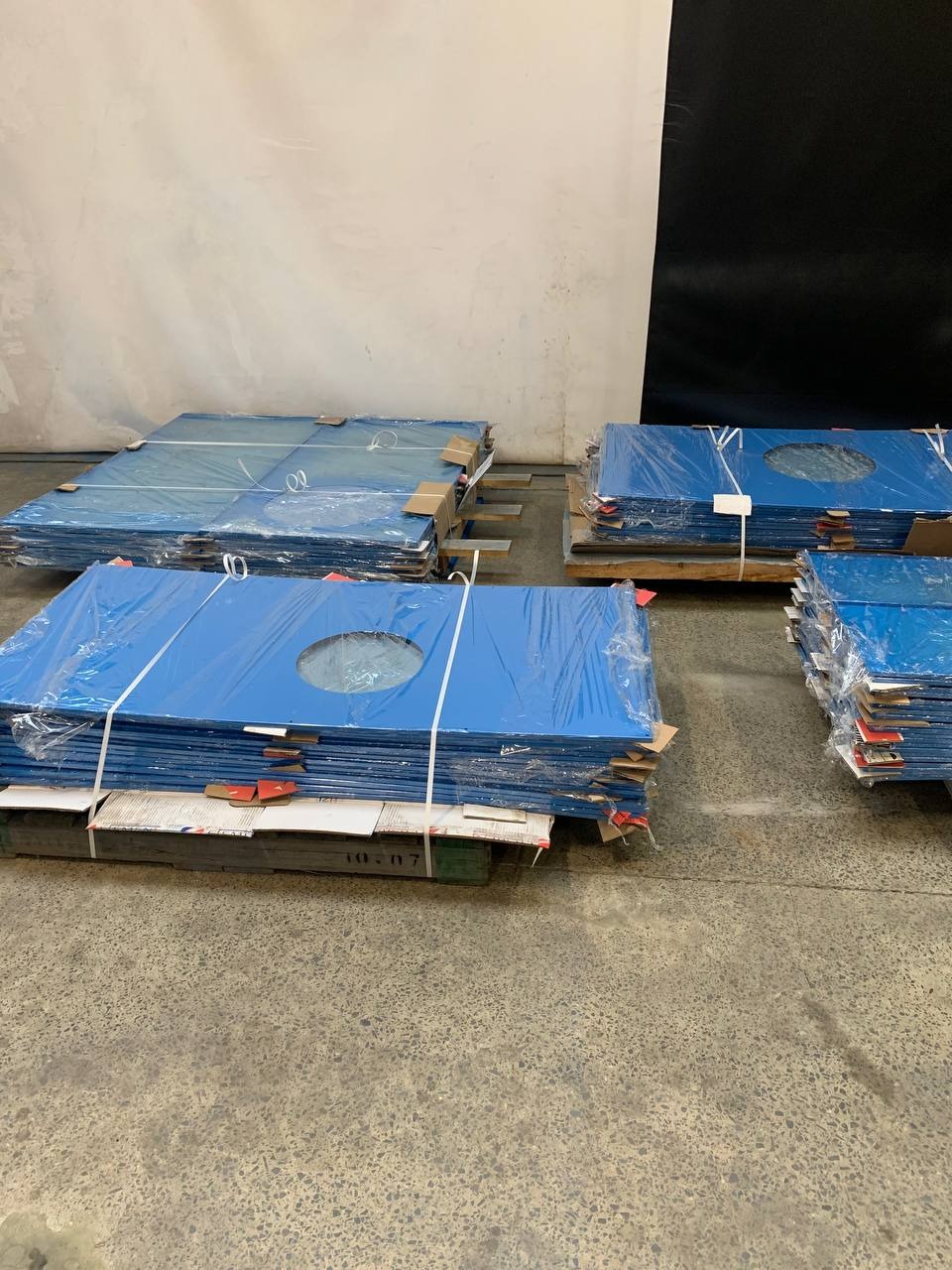 Stosy niebieskich, metalowych paneli z okrągłym otworem, zabezpieczonych folią, ułożone na paletach w hali produkcyjnej. Widoczne detale mocowań i surowy beton na podłodze.