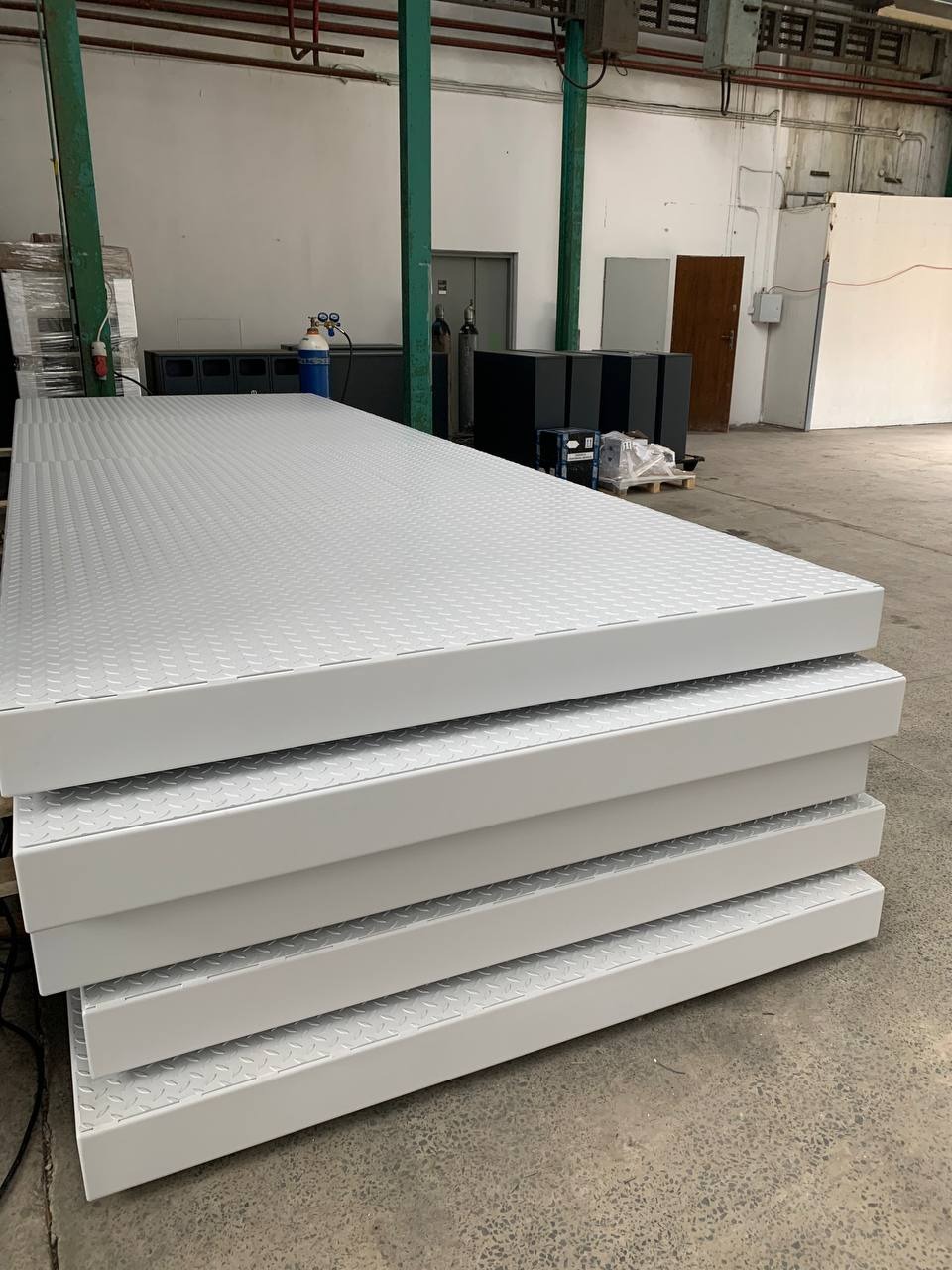 Białe, metalowe platformy z wytłaczanym wzorem, ułożone jedna na drugiej w hali produkcyjnej. Widoczne elementy konstrukcyjne i wyposażenie warsztatu w tle.