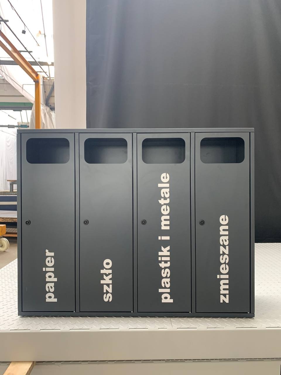 Czarny, metalowy pojemnik do segregacji odpadów (papier, szkło, plastik, zmieszane) wykonany w Błoniu. Widoczne otwory wrzutowe i napisy na frontach.