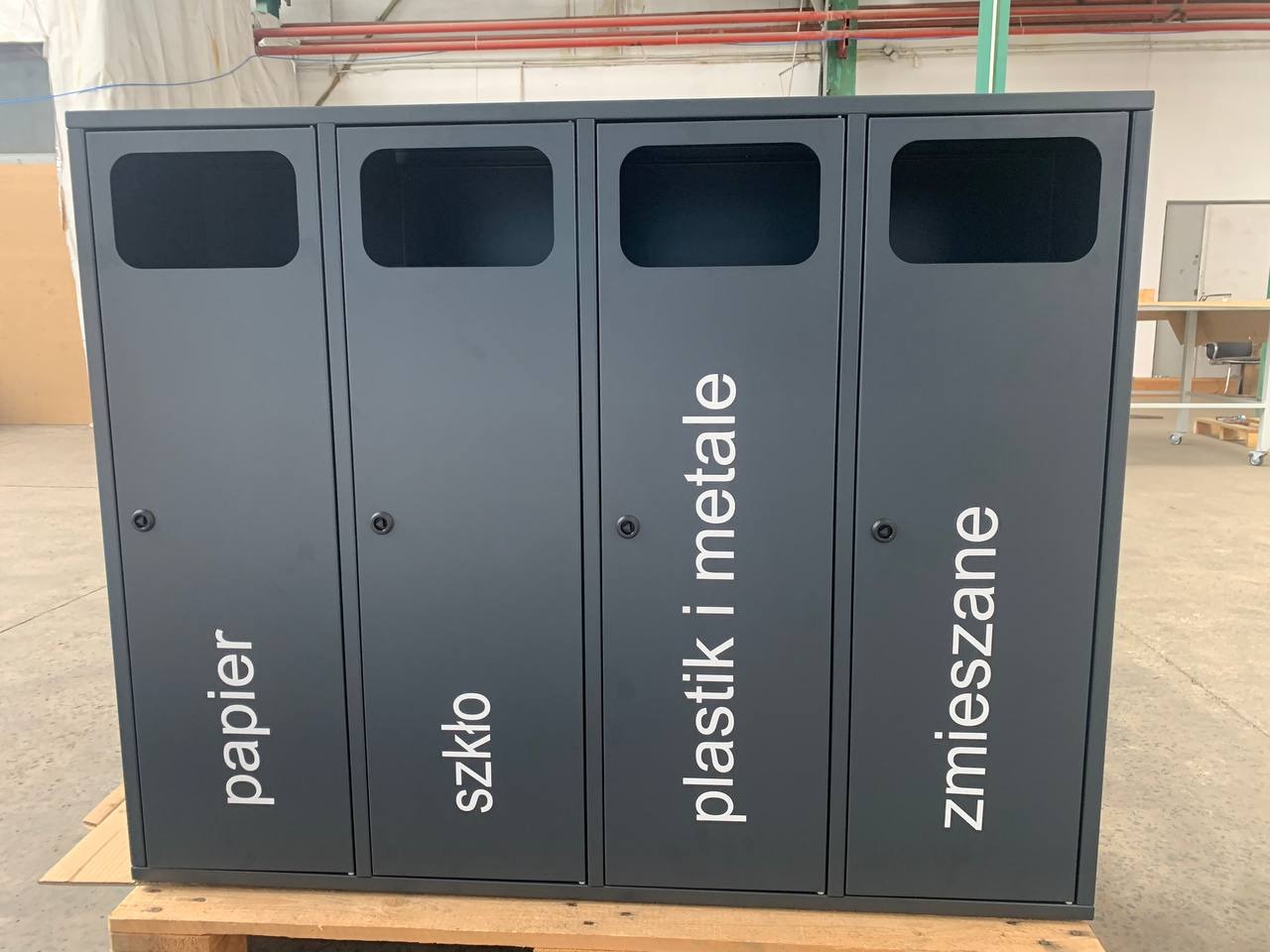 Metalowy pojemnik do segregacji odpadów z czterema przegrodami oznaczonymi: papier, szkło, plastik i metale, zmieszane. Szara, matowa powierzchnia, widoczne zamki.
